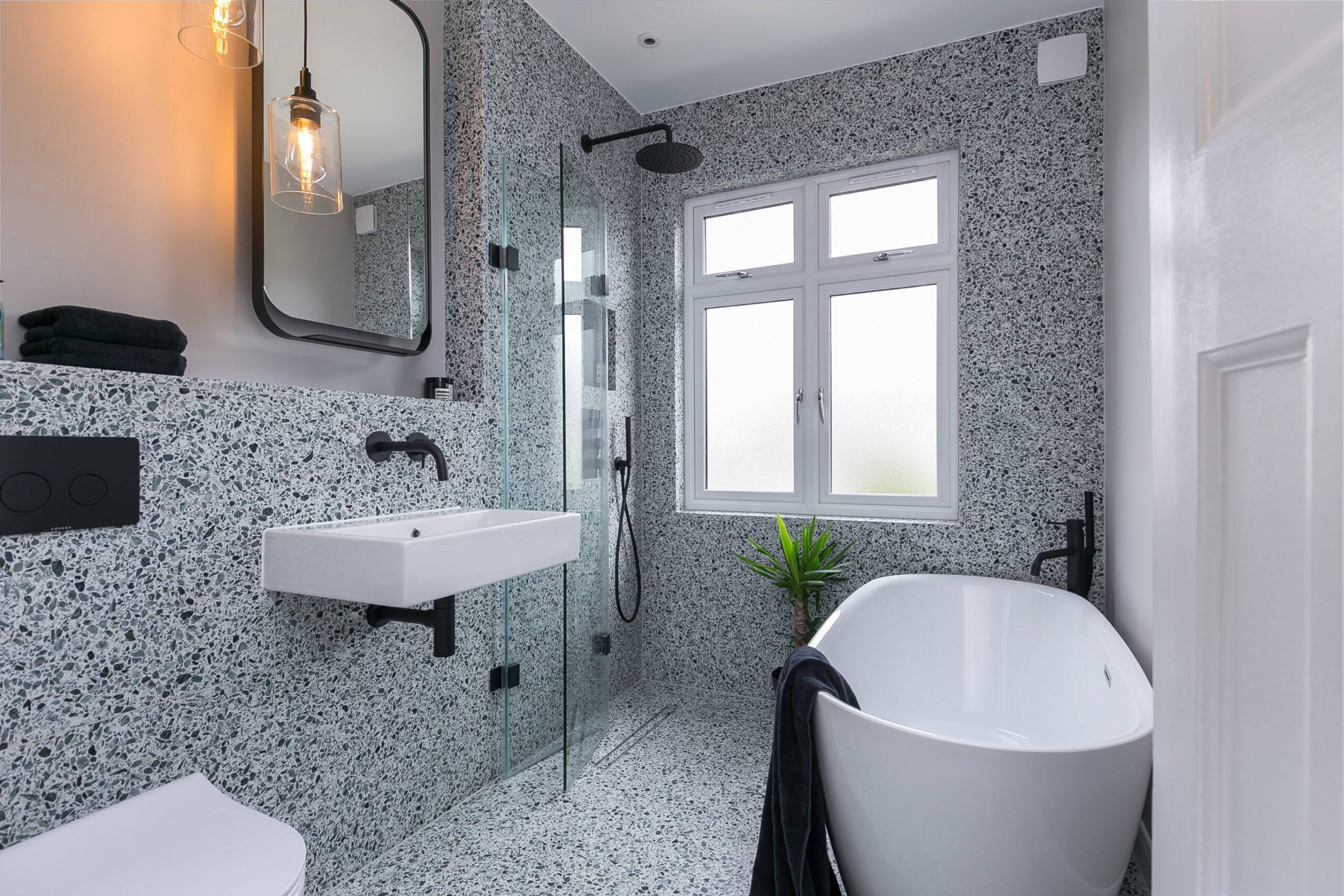 Modern bathroom featuring terrazzo walls and flooring, a frameless glass shower, white freestanding bathtub, black fixtures, and a large window.