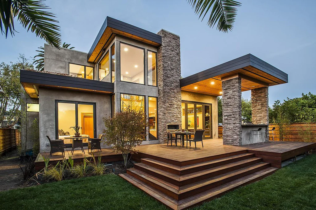 Contemporary two-story prefab home with large glass windows, stone columns, and a spacious wooden deck featuring outdoor dining and a built-in bar area.