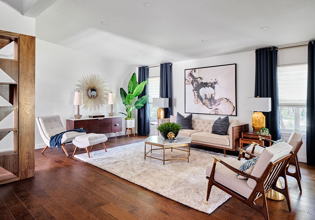 Stylish living room featuring a beige tufted sofa, wood accent chairs, a large abstract art piece, starburst mirror, and dark drapes on tall windows.