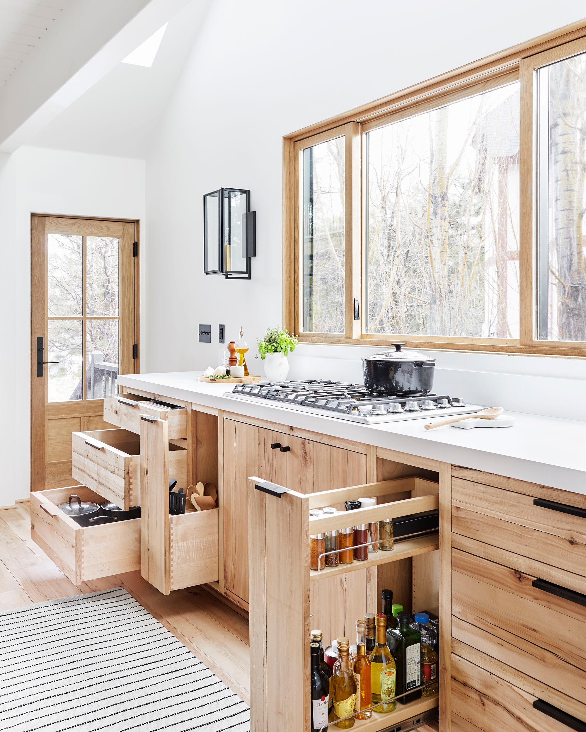 Minimalist kitchen featuring light natural wood cabinets with pull-out drawers, a white countertop, large windows, and organized spice storage.