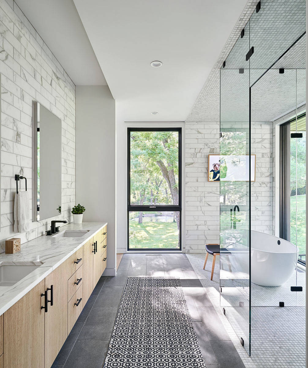 Modern bathroom with glass shower, freestanding tub, marble walls, and large window overlooking greenery.