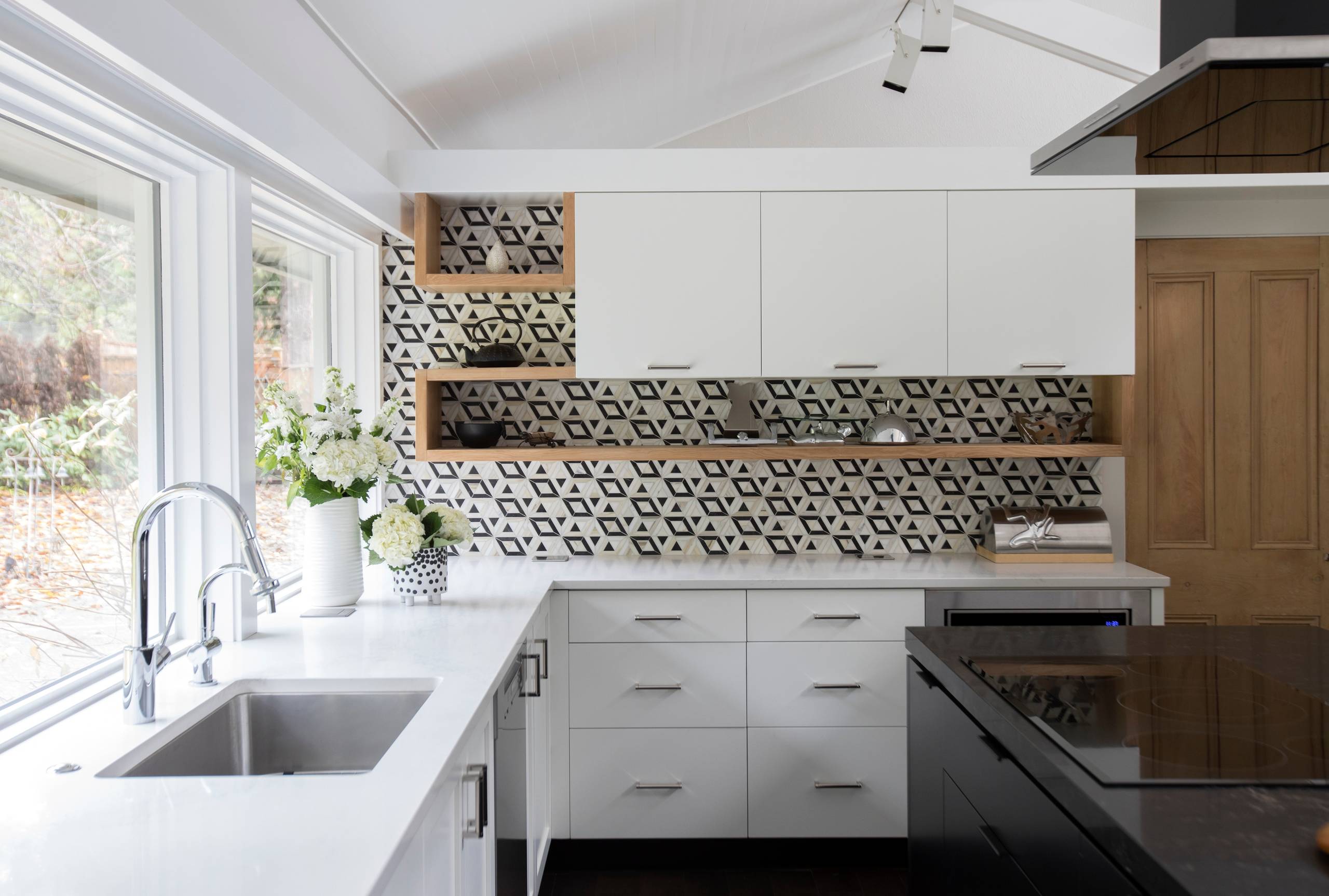 Contemporary kitchen featuring white cabinets, a geometric black and white tile backsplash, built-in wooden shelving, and a sleek island cooktop.