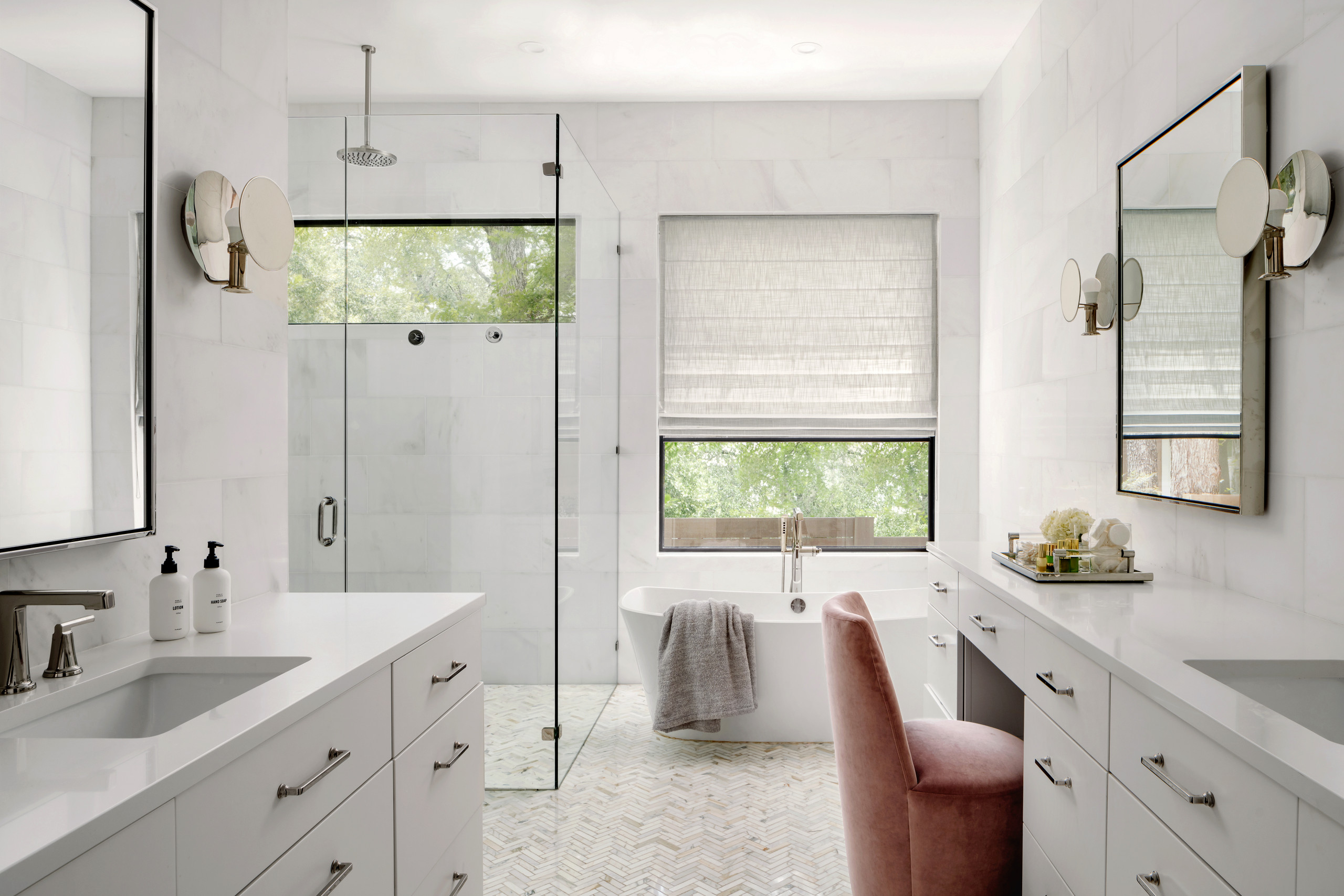 Elegant modern bathroom with freestanding tub, glass shower, double vanity, and pink accent chair.