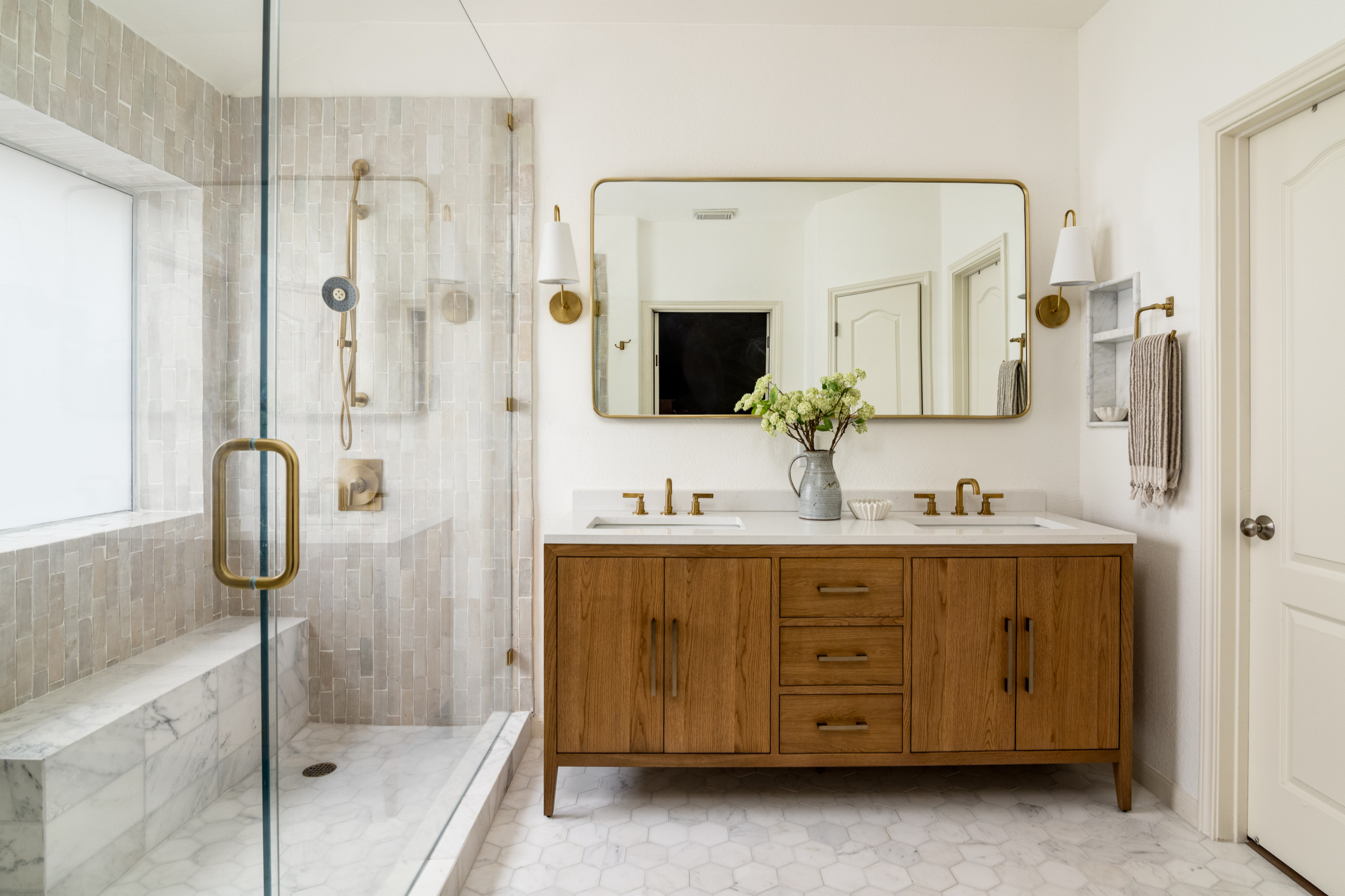 Elegant bathroom with a wooden double vanity, gold fixtures, glass shower, and marble tile accents.