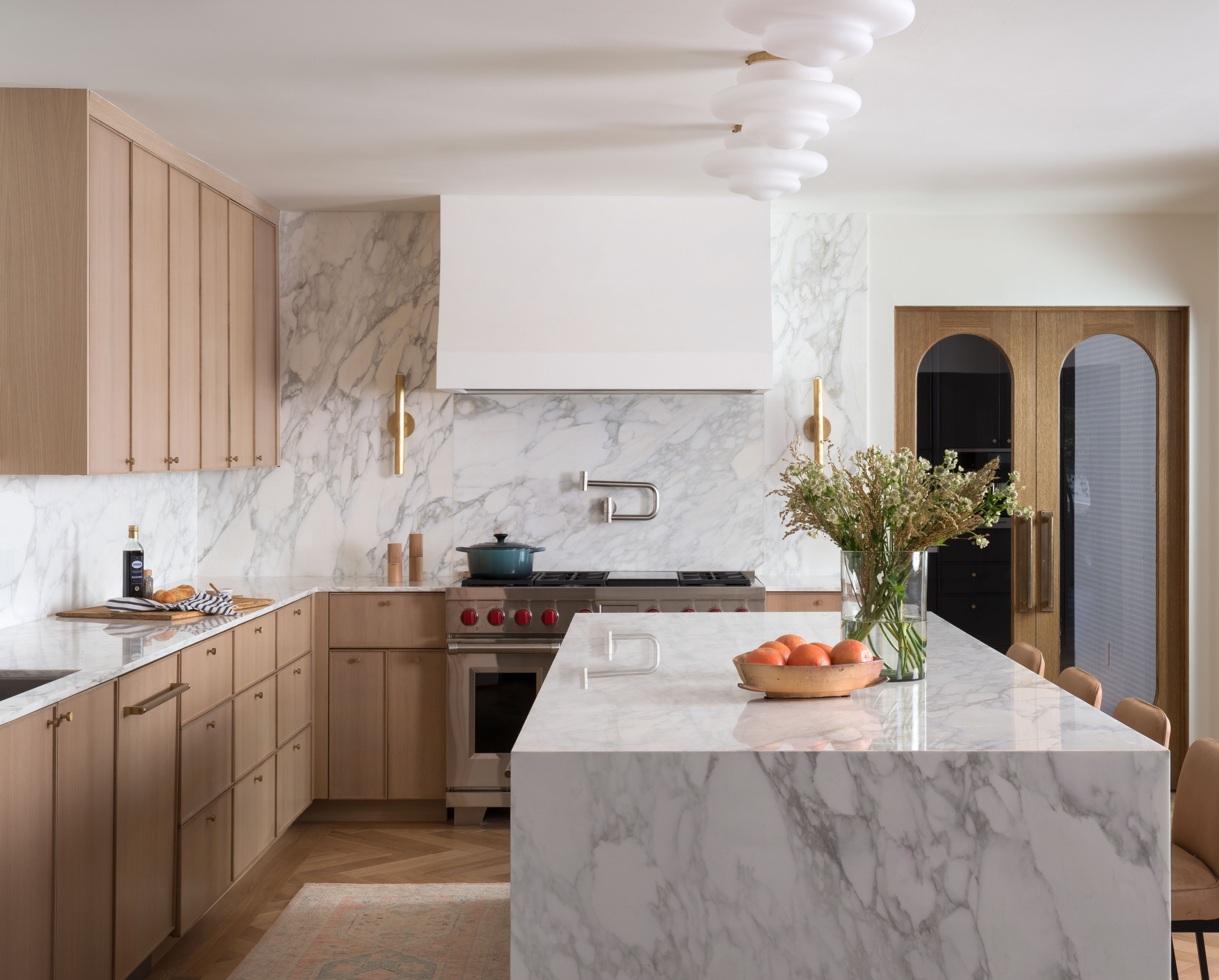 Modern kitchen with marble countertops and backsplash, light wood cabinetry, and a central island decorated with flowers and oranges.