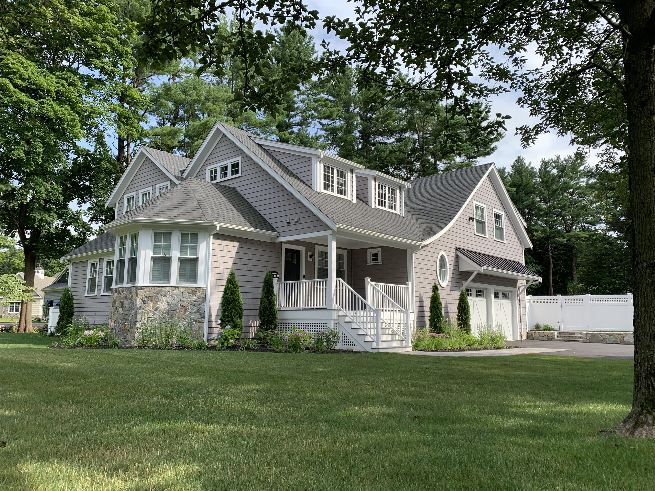 Custom-built two-story suburban home with gray siding, stone accents, white trim, and a front porch surrounded by greenery.