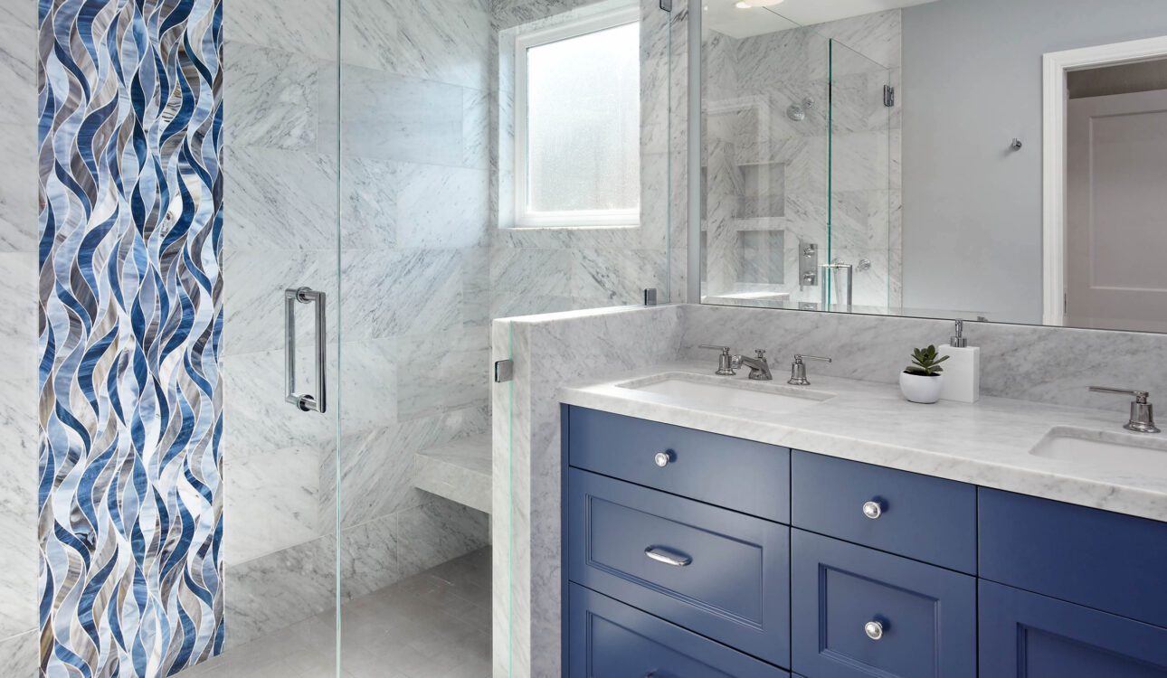 Bathroom with blue vanity, marble countertops, and glass shower featuring decorative blue tile accent.