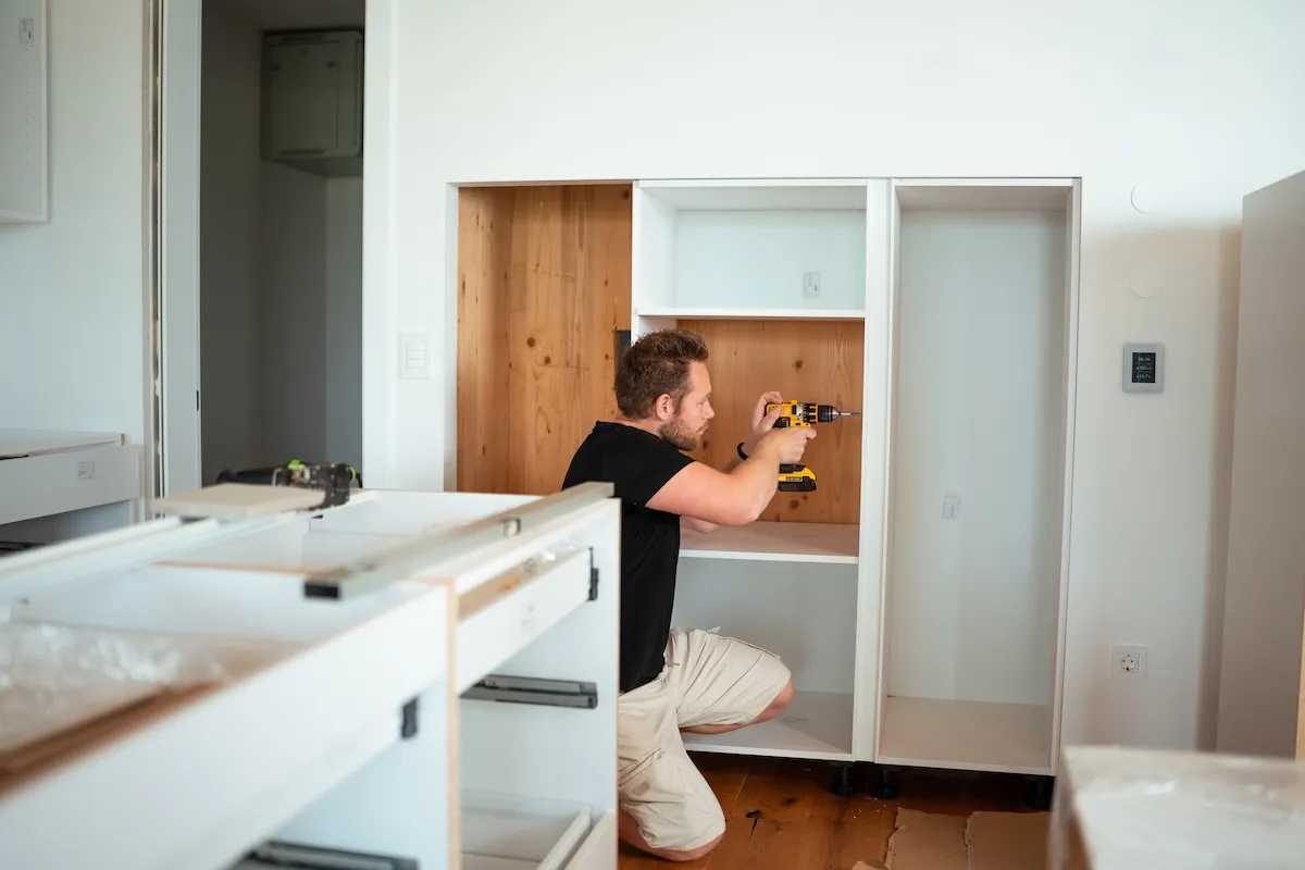 A man kneeling and using a power drill to install a cabinet shelf.