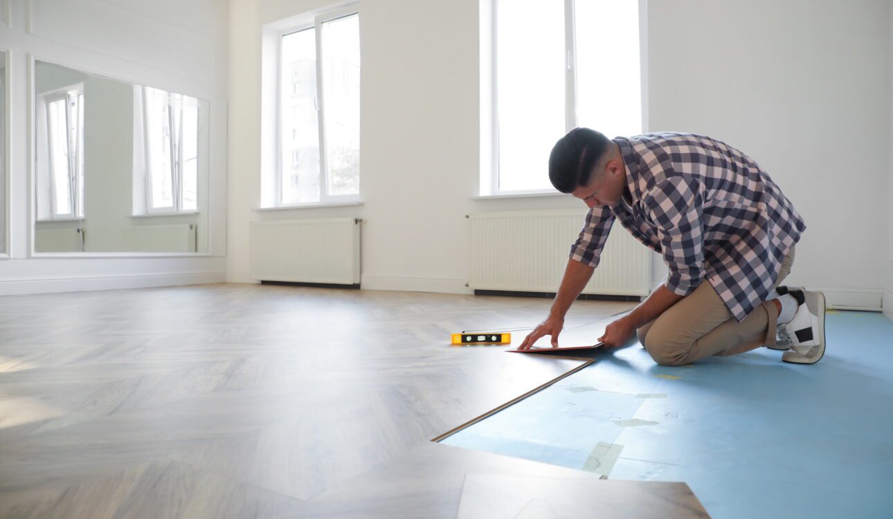 Man installing vinyl plank flooring over a blue underlayment in a bright modern room.