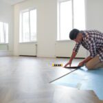 Man installing vinyl plank flooring over a blue underlayment in a bright modern room.