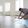 Man installing vinyl plank flooring over a blue underlayment in a bright modern room.