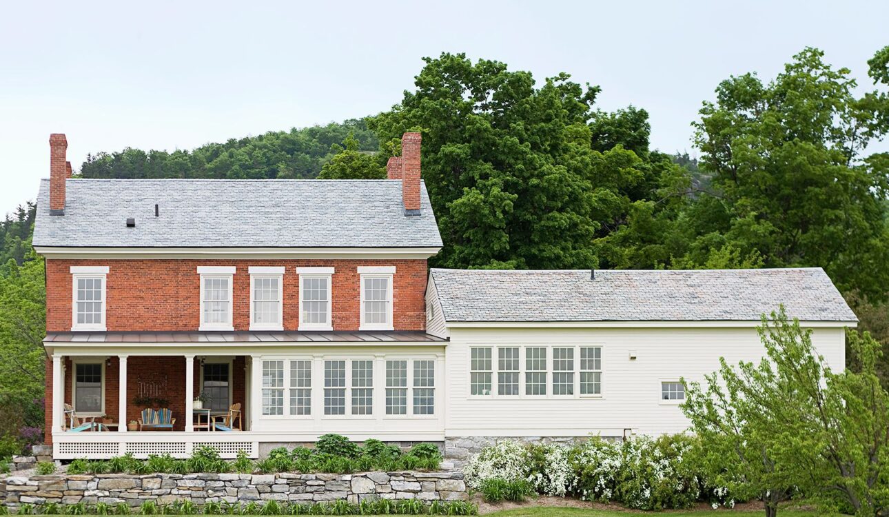Traditional brick home with a white siding addition, large windows, and a spacious front porch surrounded by greenery.