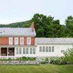 Traditional brick home with a white siding addition, large windows, and a spacious front porch surrounded by greenery.