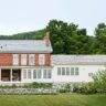 Traditional brick home with a white siding addition, large windows, and a spacious front porch surrounded by greenery.